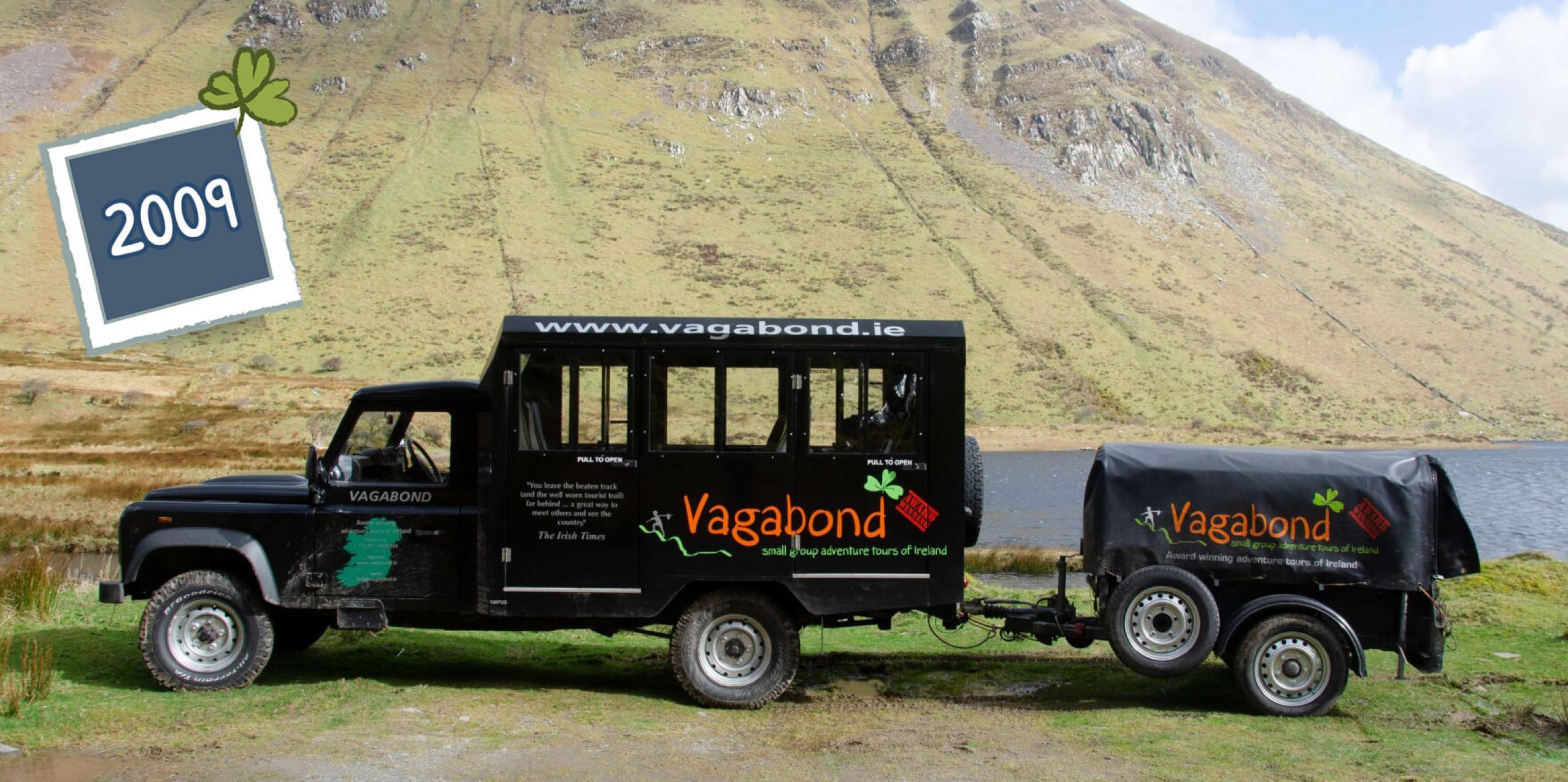 An original Land Rover Defender VagaTron tour vehicle with branded trailer parked in a scenic location in Ireland; side view
