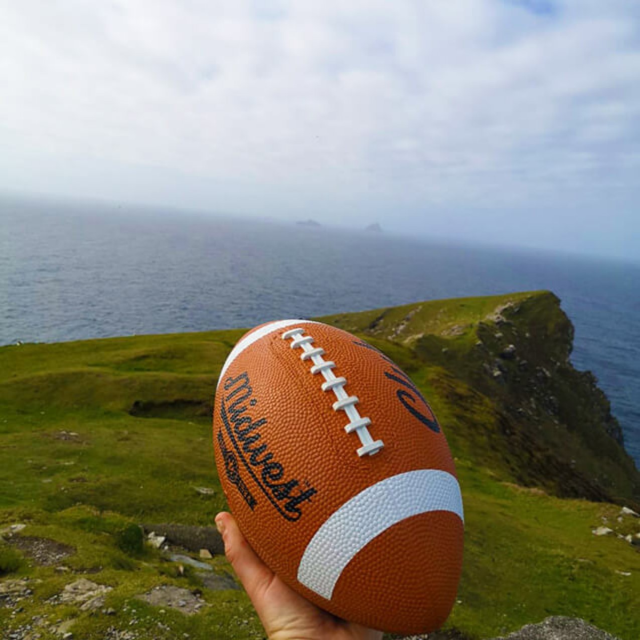 A hand holding an American football on Bray head with a view of the Skellig Islands in the background