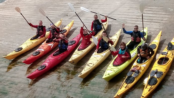 Group of Vagabond guests waving their paddles while sitting in kayaks on a slipway